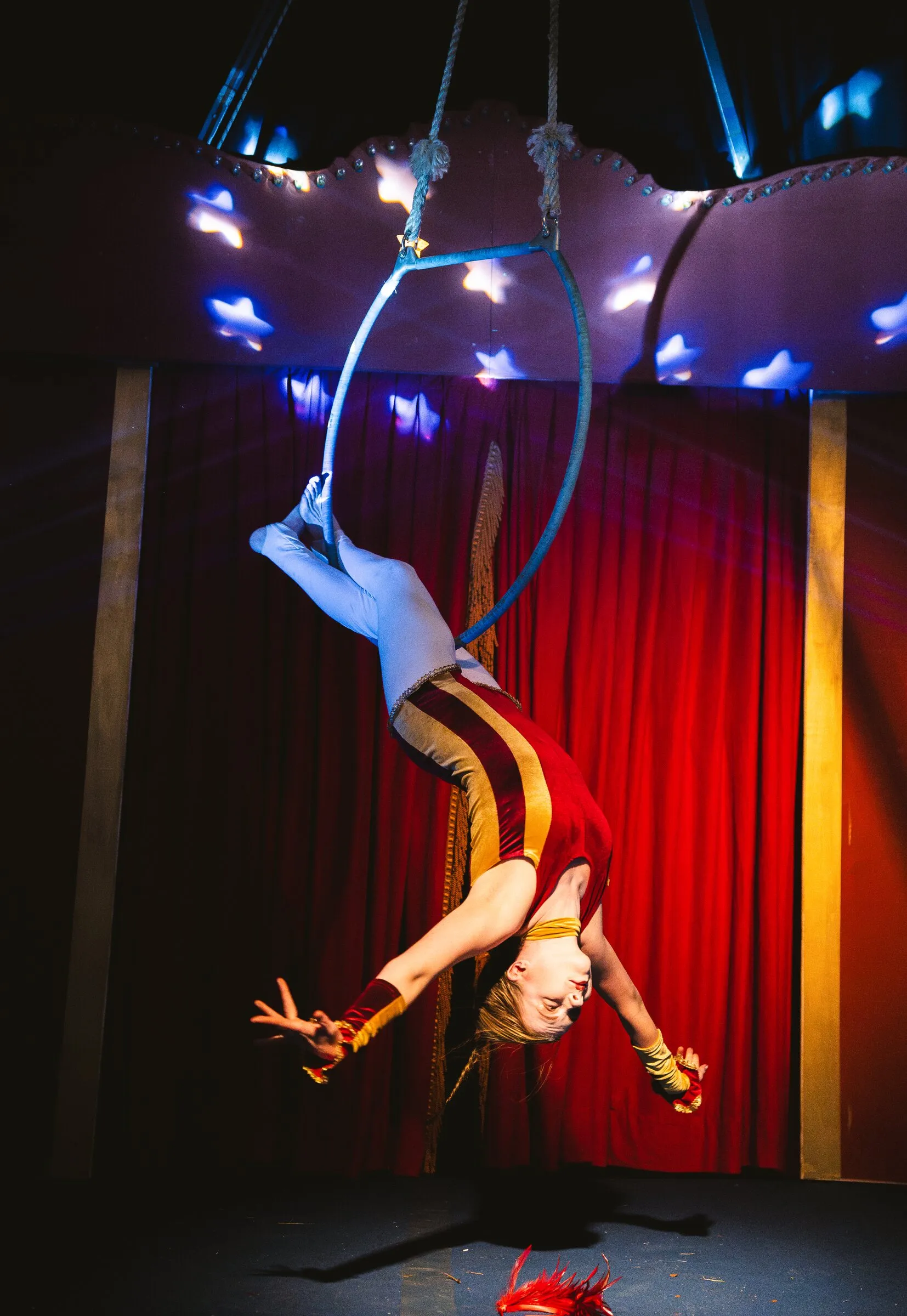 A Kuroneko Circus aerialist hanging backward from an aerial hoop beneath lit stars