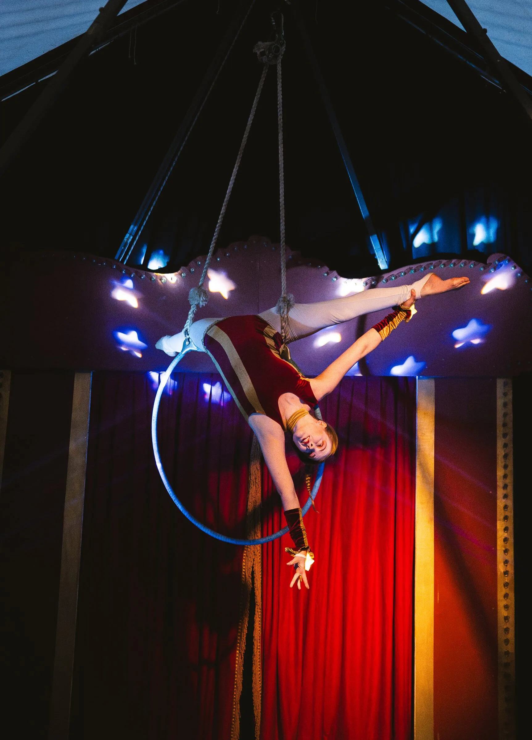 A Kuroneko Circus aerialist stretched in a layout beneath an aerial hoop under the star canopy