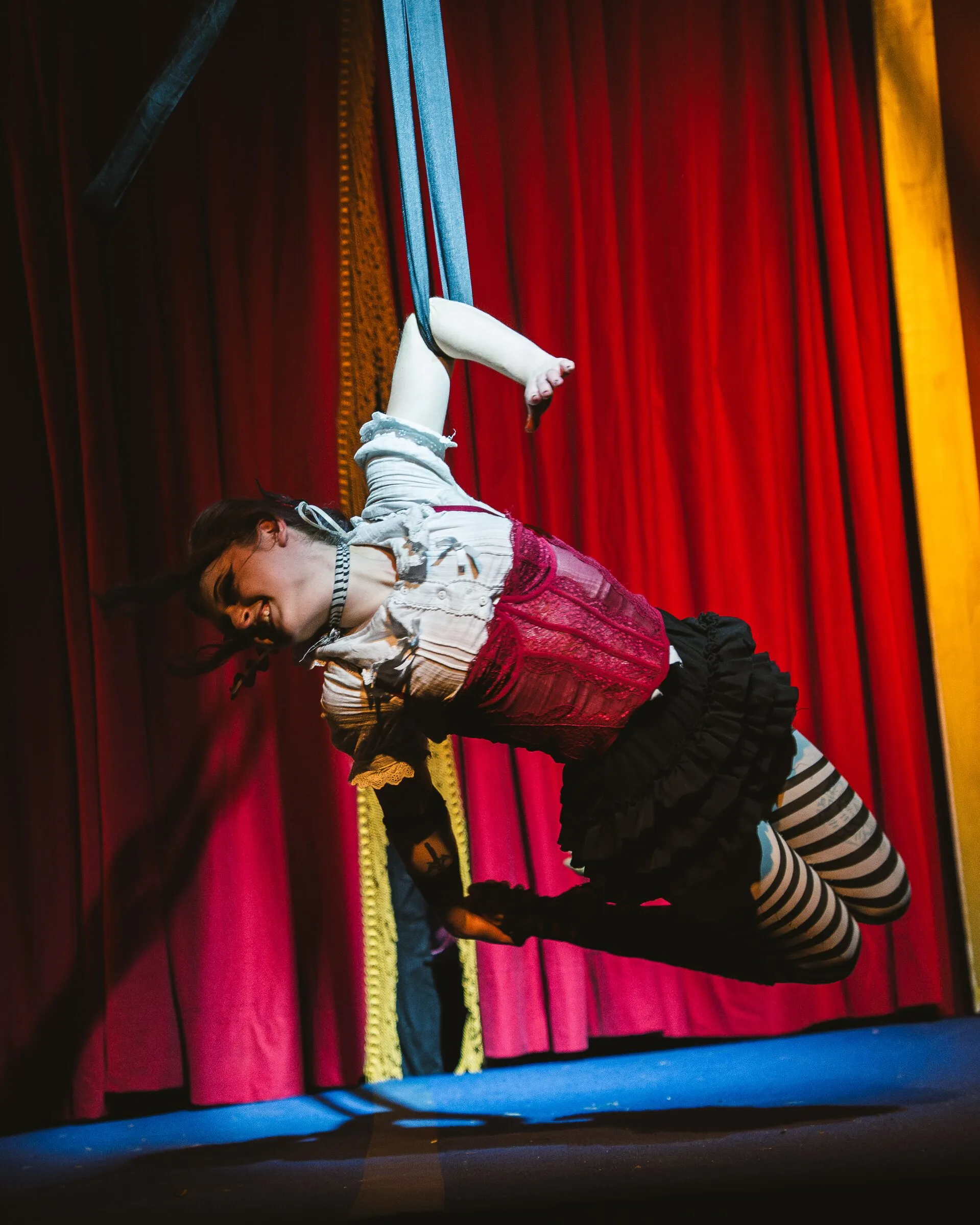 A Kuroneko Circus aerialist in striped stockings diving forward from aerial loops above the stage