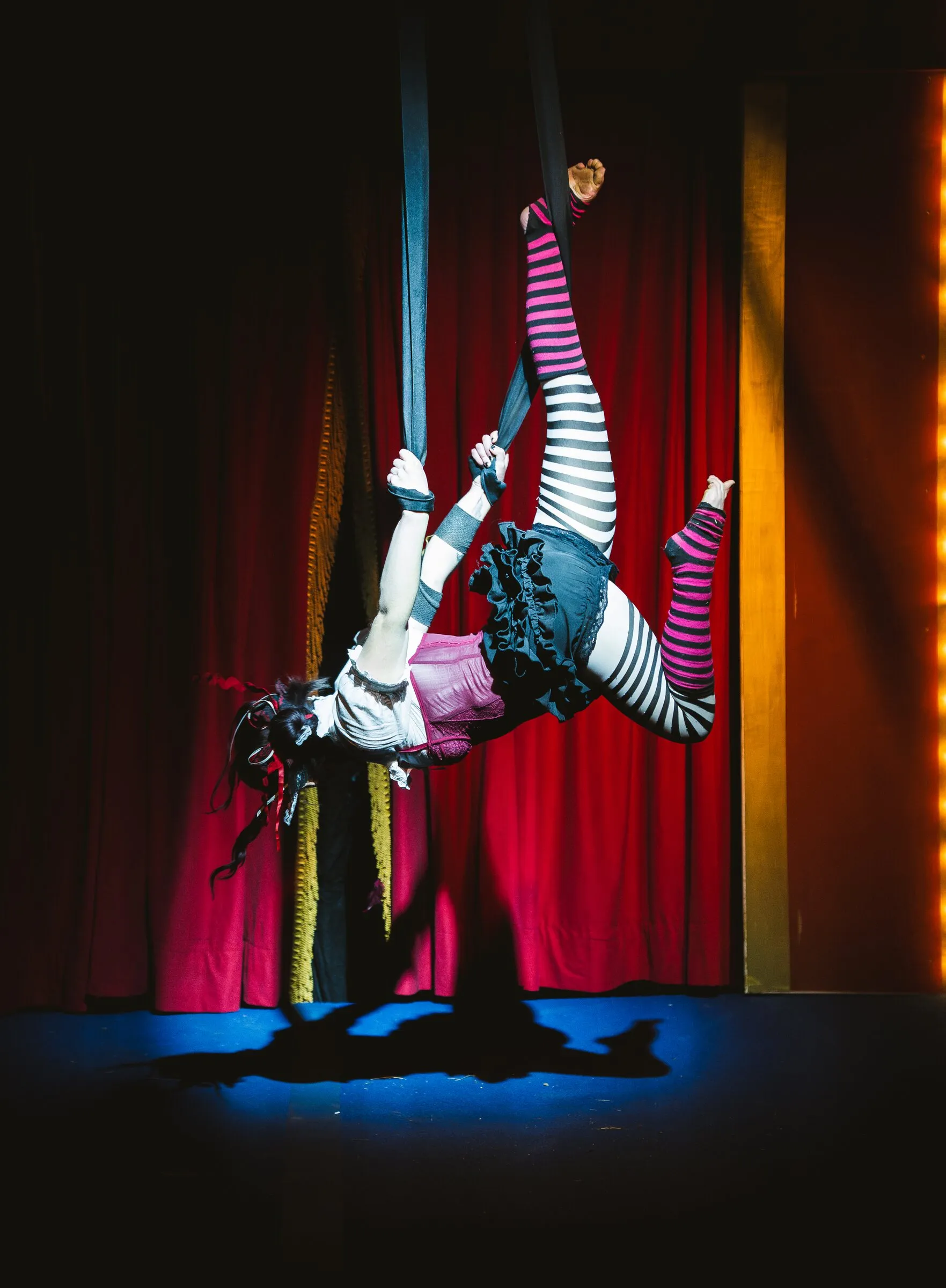 A Kuroneko Circus aerialist inverted in aerial loops above a blue-lit stage