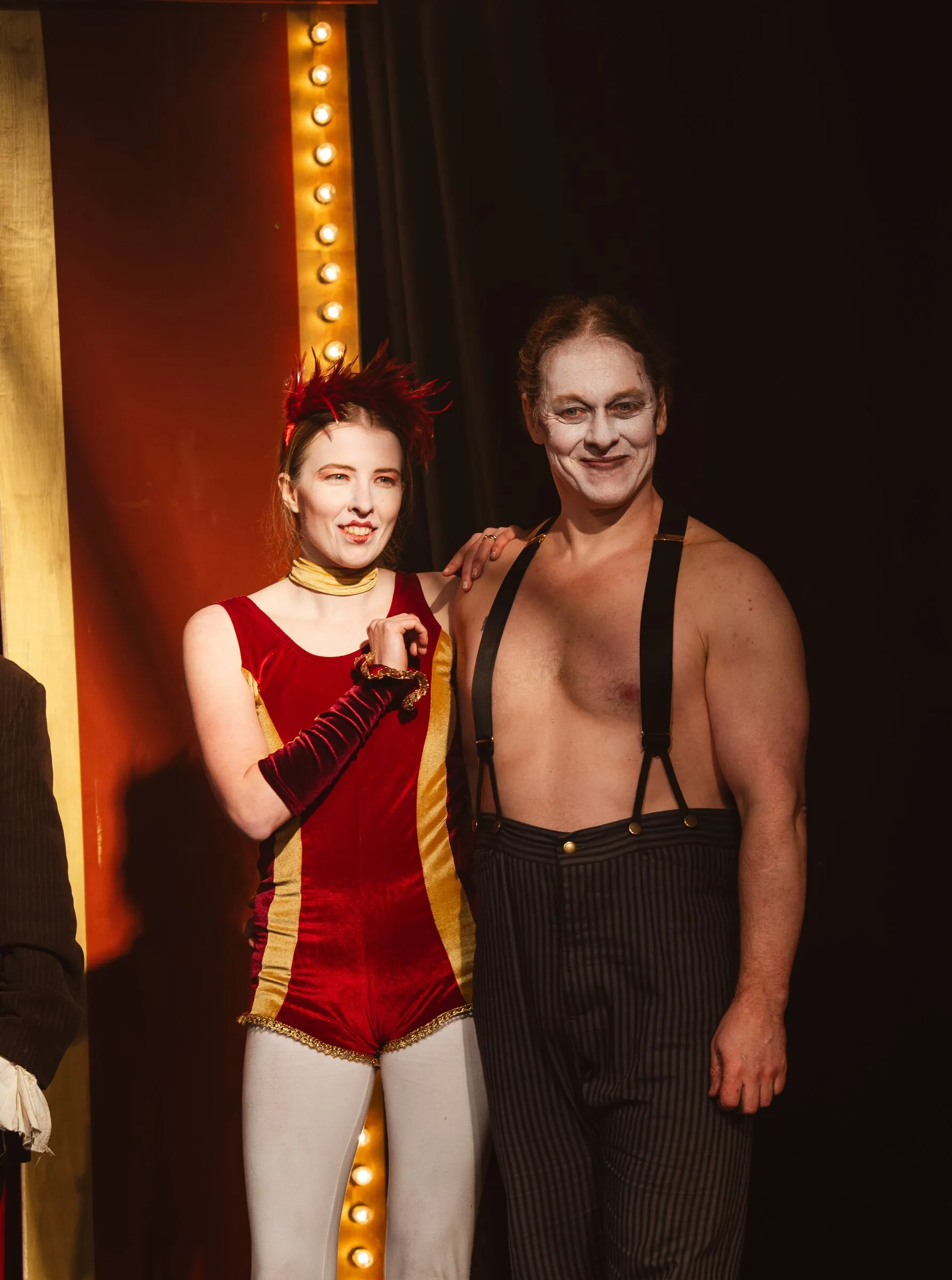 A Kuroneko Circus aerialist and strongman smiling together beside the marquee lights after the show