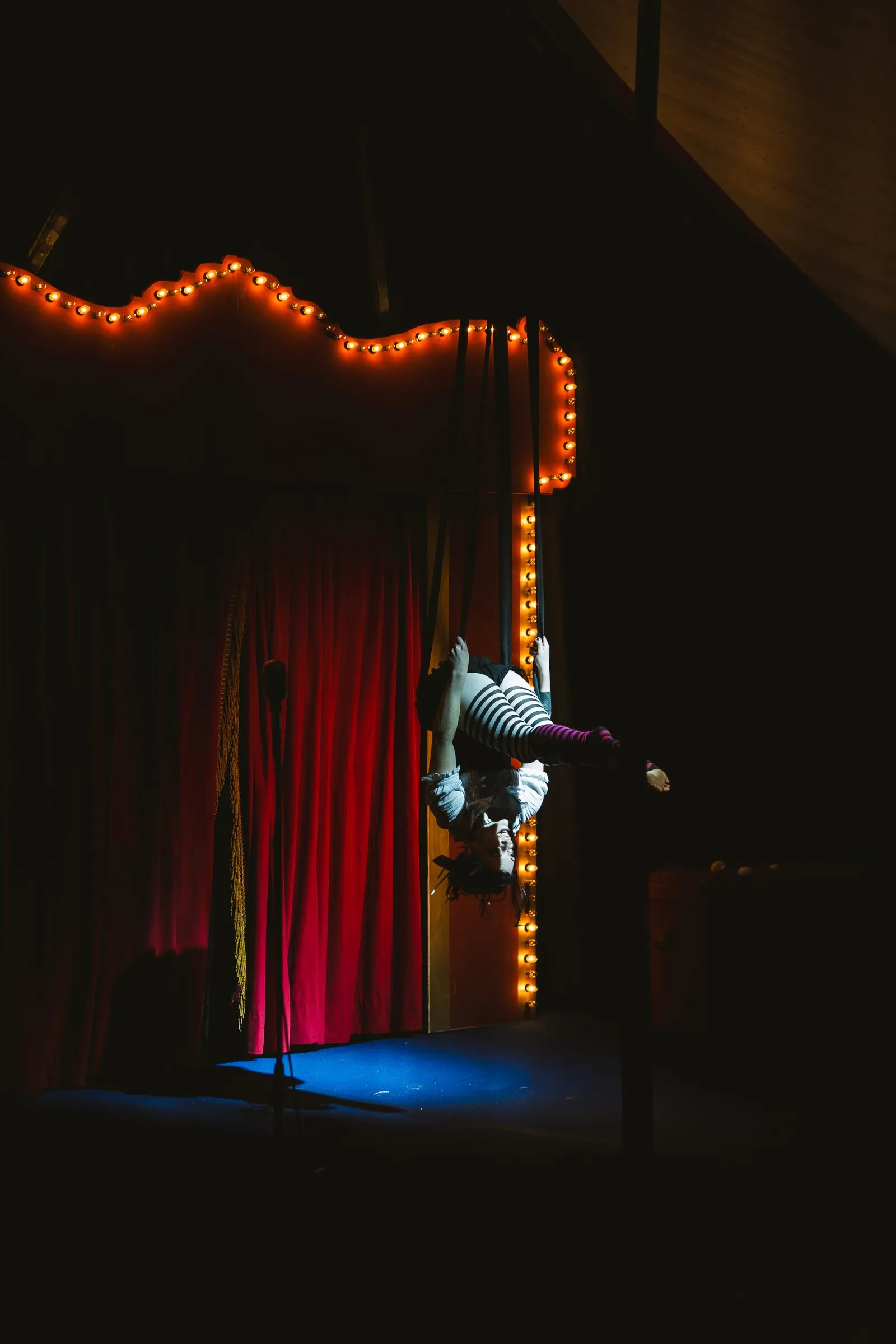 A Kuroneko Circus aerialist in striped stockings hanging upside down beside the lit marquee arch