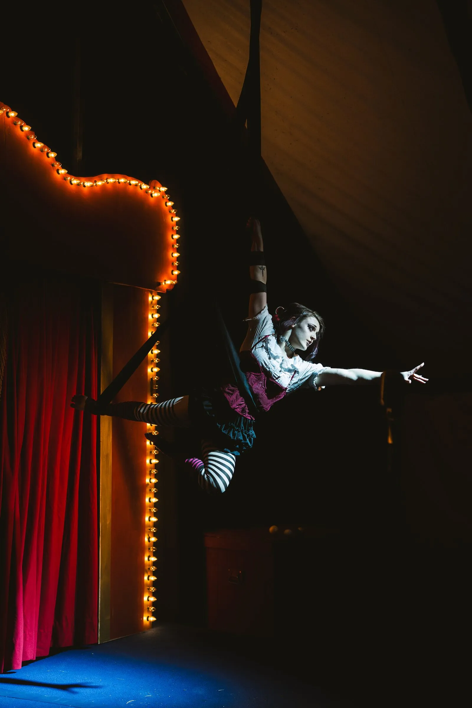 A Kuroneko Circus aerialist in striped stockings reaching out from aerial loops beside the marquee lights