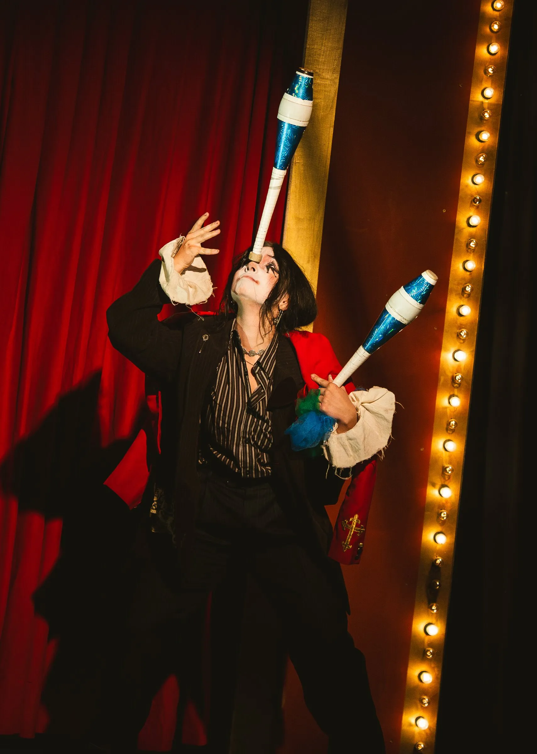 A Kuroneko Circus clown in whiteface balancing a club on her face beside the marquee lights