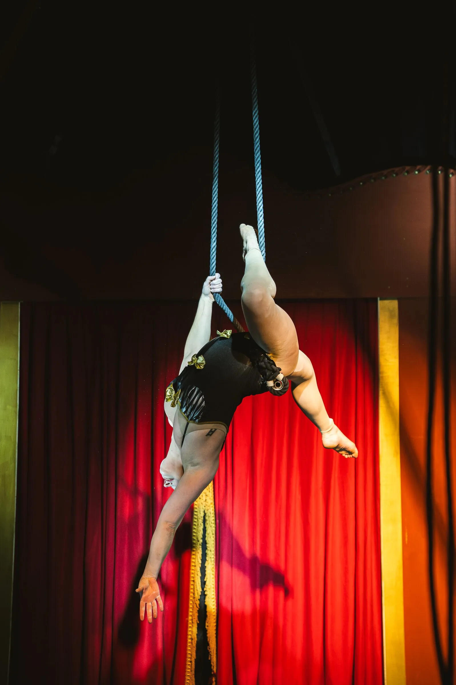 A Kuroneko Circus trapeze artist hanging from the dance trapeze, reaching down toward the stage