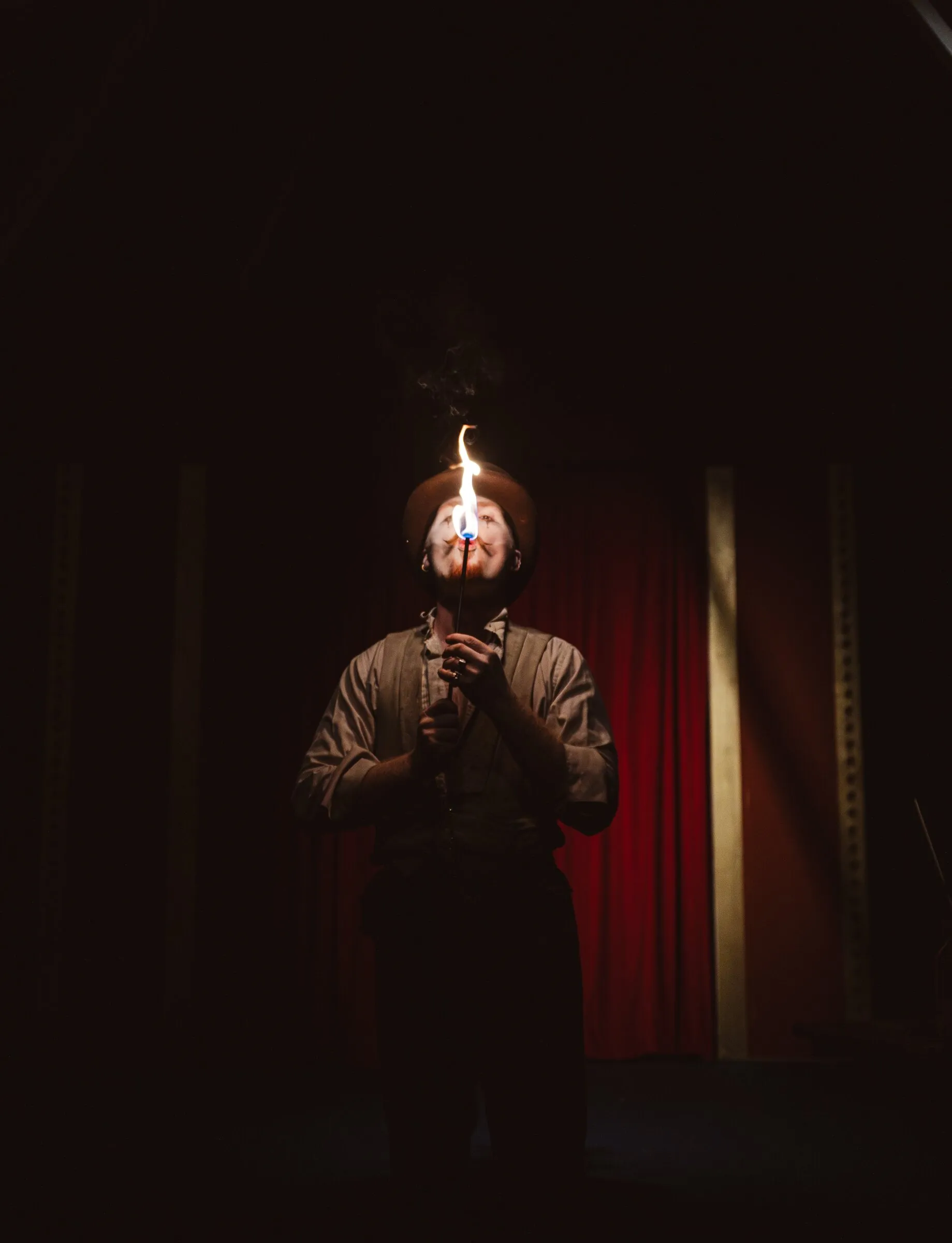 A Kuroneko Circus fire eater lighting a torch close to his face in a dark tent