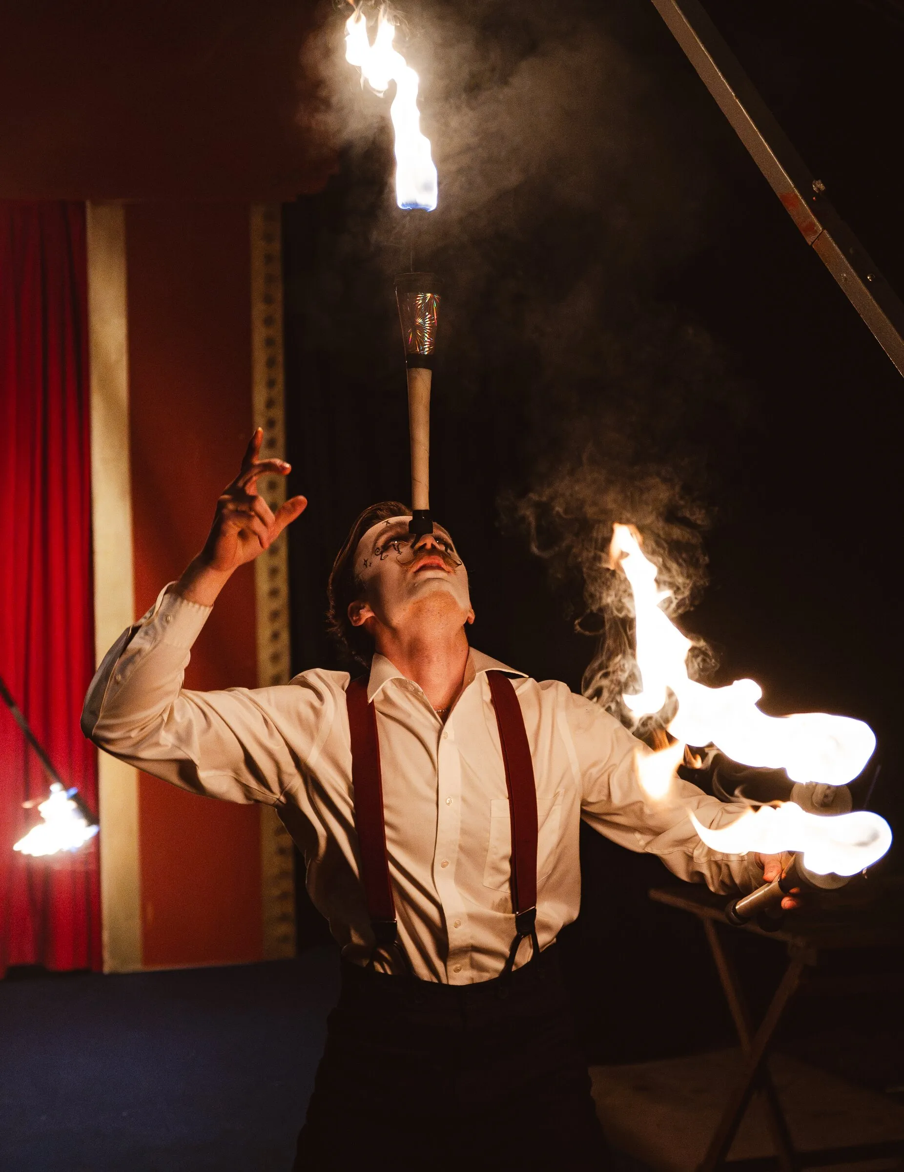 A Kuroneko Circus performer in suspenders balancing a flaming torch on his chin while juggling fire under the big top in North Carolina