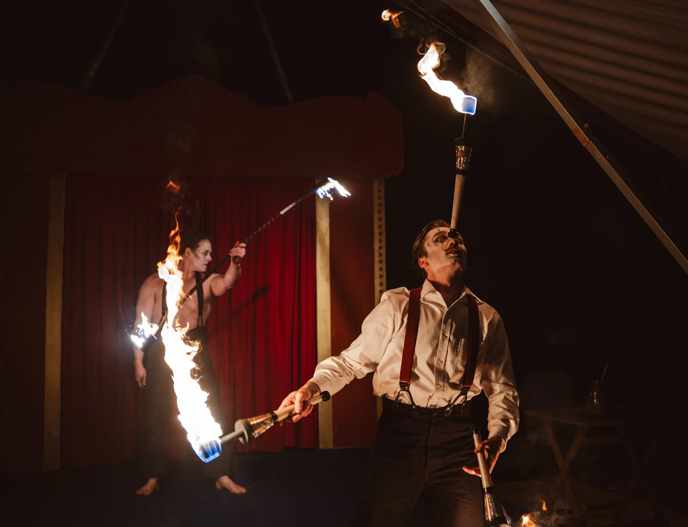 A Kuroneko Circus juggler balancing a flaming torch on his chin while a partner spins fire behind him