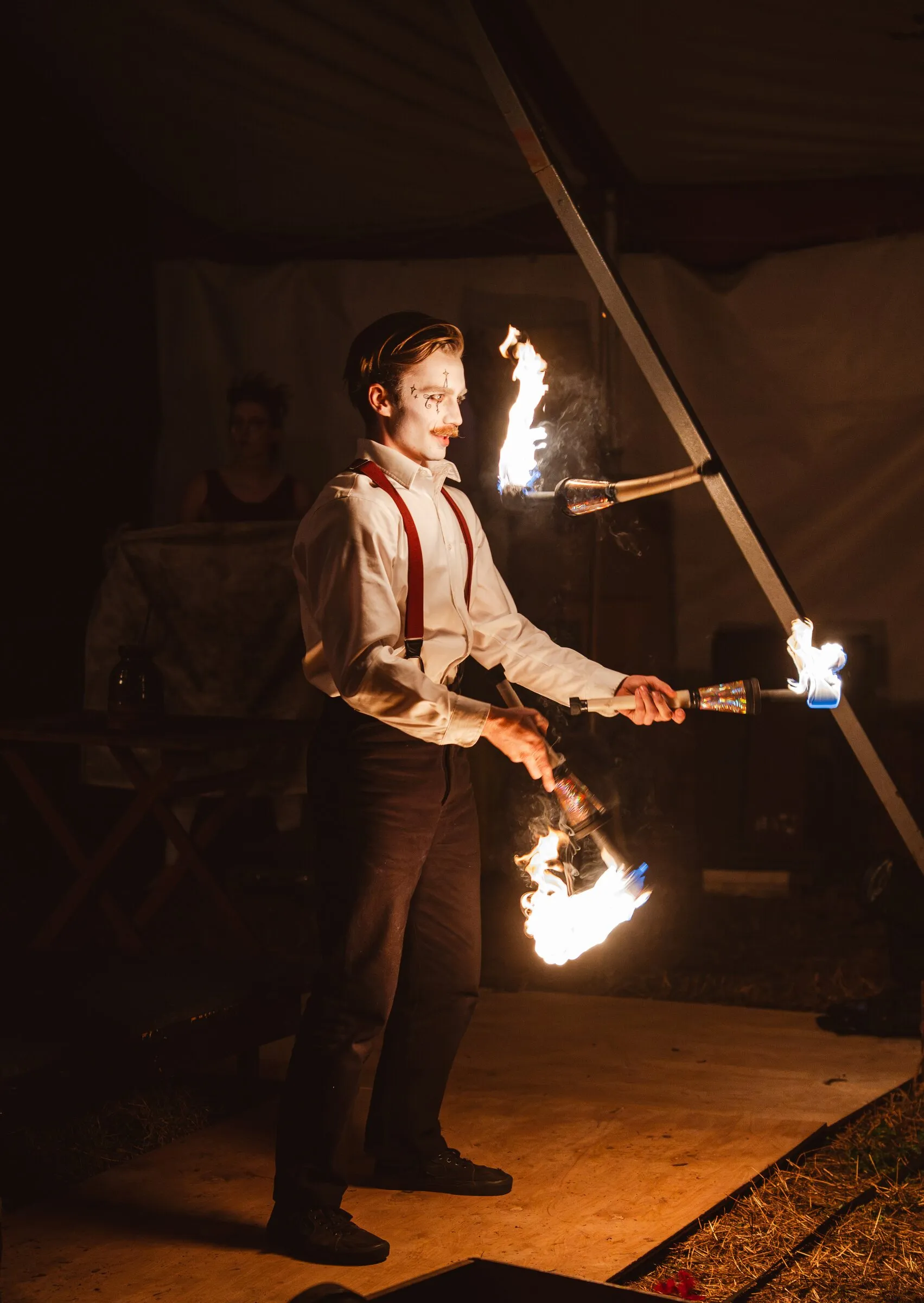 A Kuroneko Circus juggler in suspenders juggling flaming torches on the wooden stage