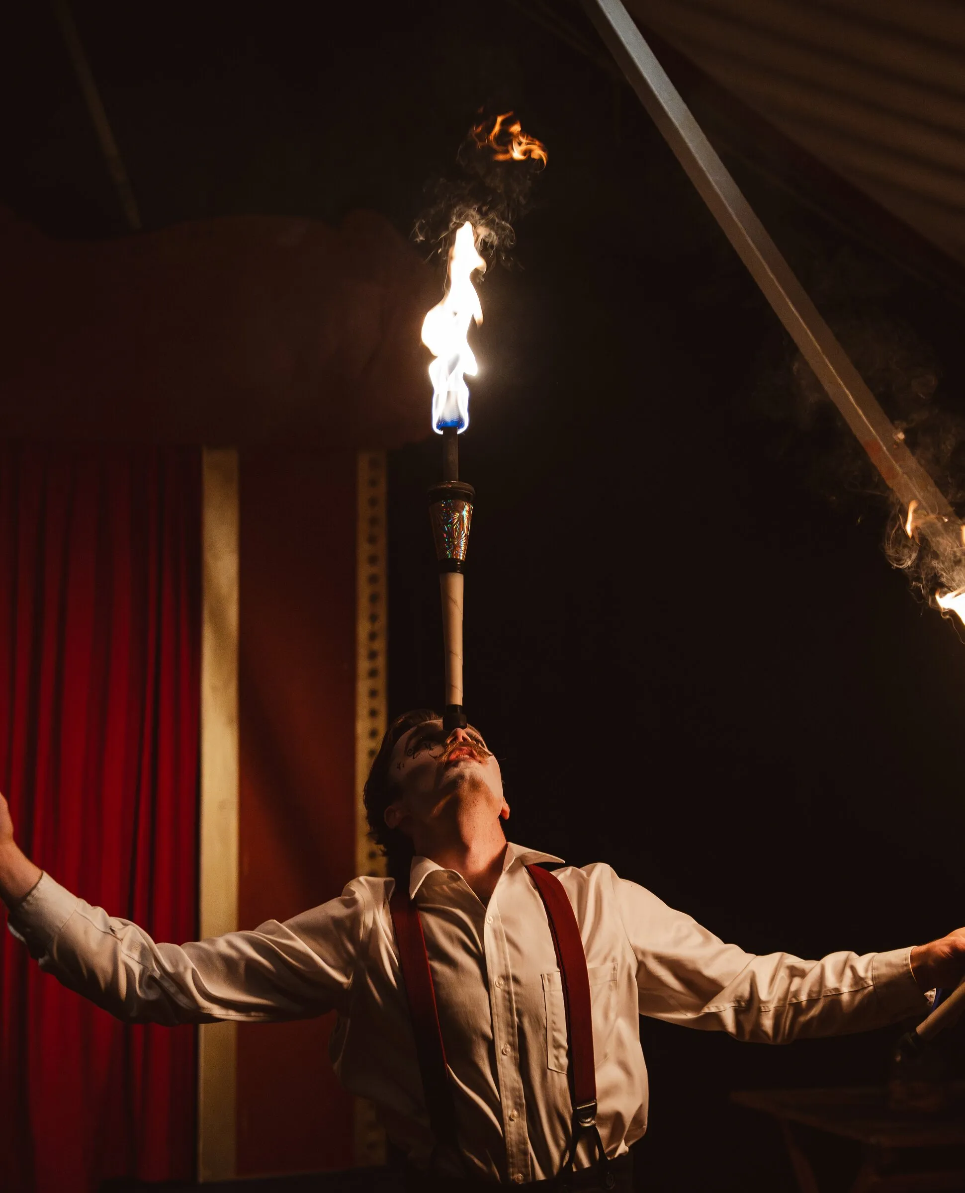 A Kuroneko Circus fire performer balancing a flaming torch on his chin with his arms thrown wide