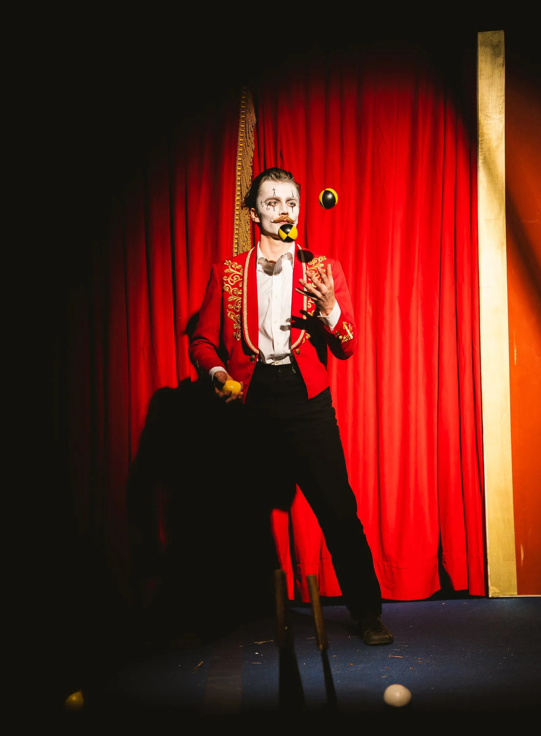 A Kuroneko Circus juggler in whiteface and a red tailcoat keeping balls in the air in a spotlight