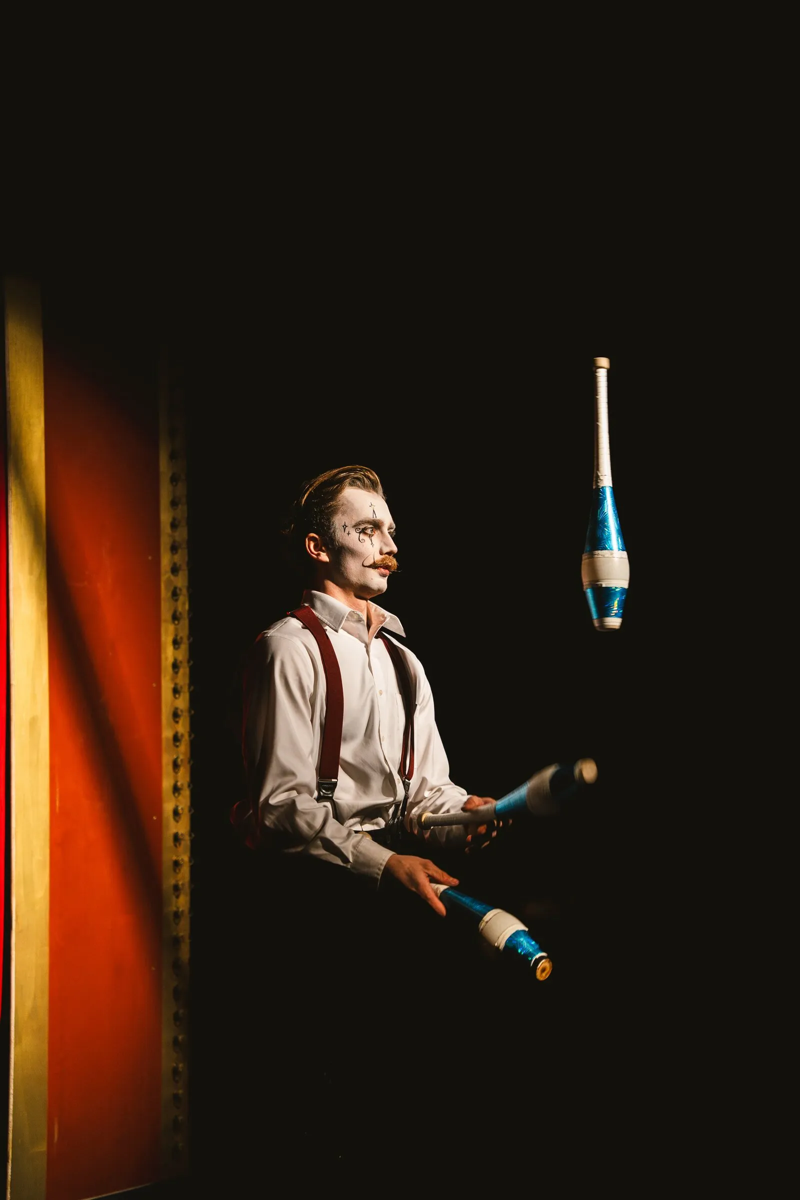 A Kuroneko Circus juggler in whiteface and suspenders juggling clubs in a narrow spotlight
