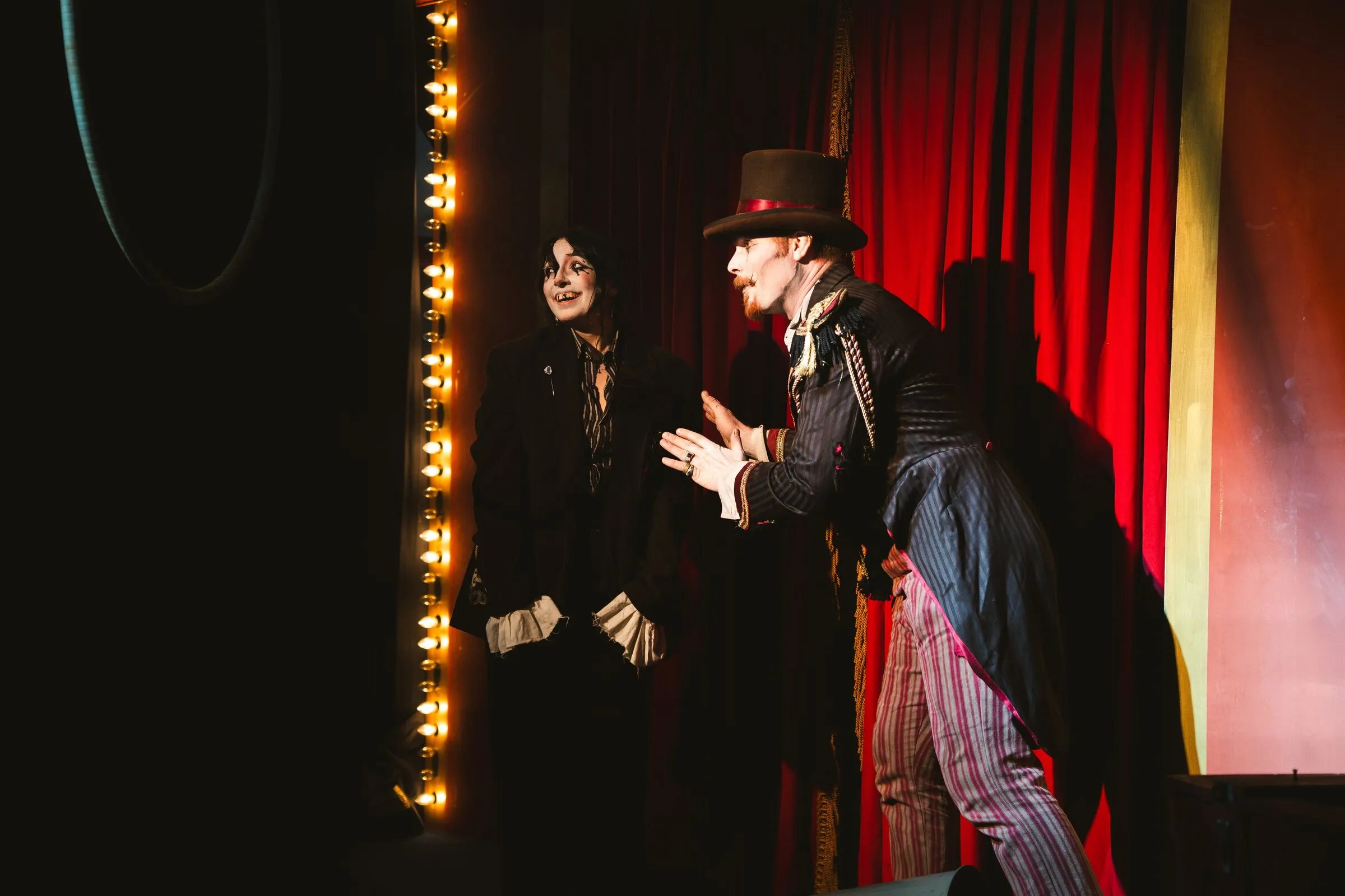 The Kuroneko Circus ringmaster leaning in toward a sideshow clown beside the marquee lights
