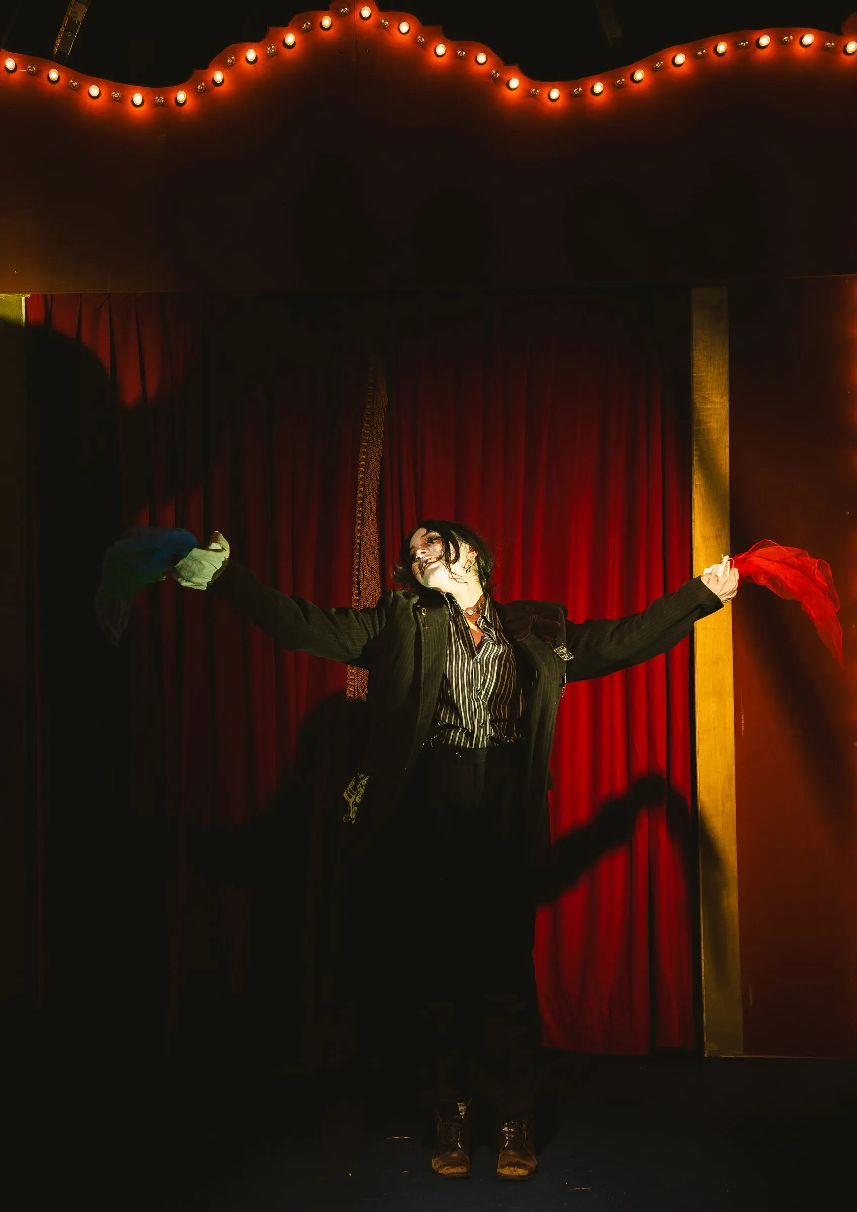 A Kuroneko Circus sideshow clown in whiteface flinging bright scarves wide before the red curtain