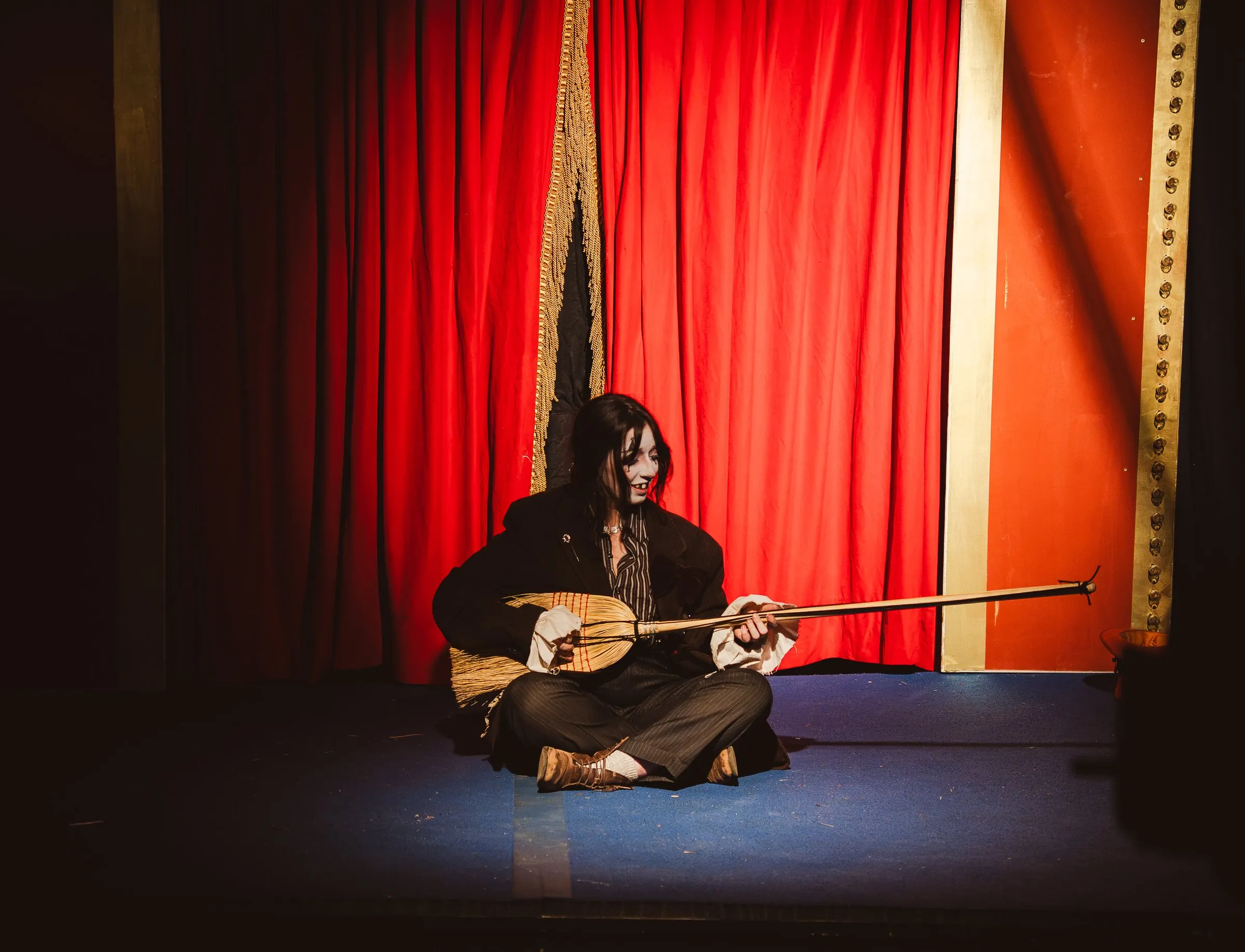 A Kuroneko Circus sideshow clown sitting cross-legged on the stage playing a long-necked lute