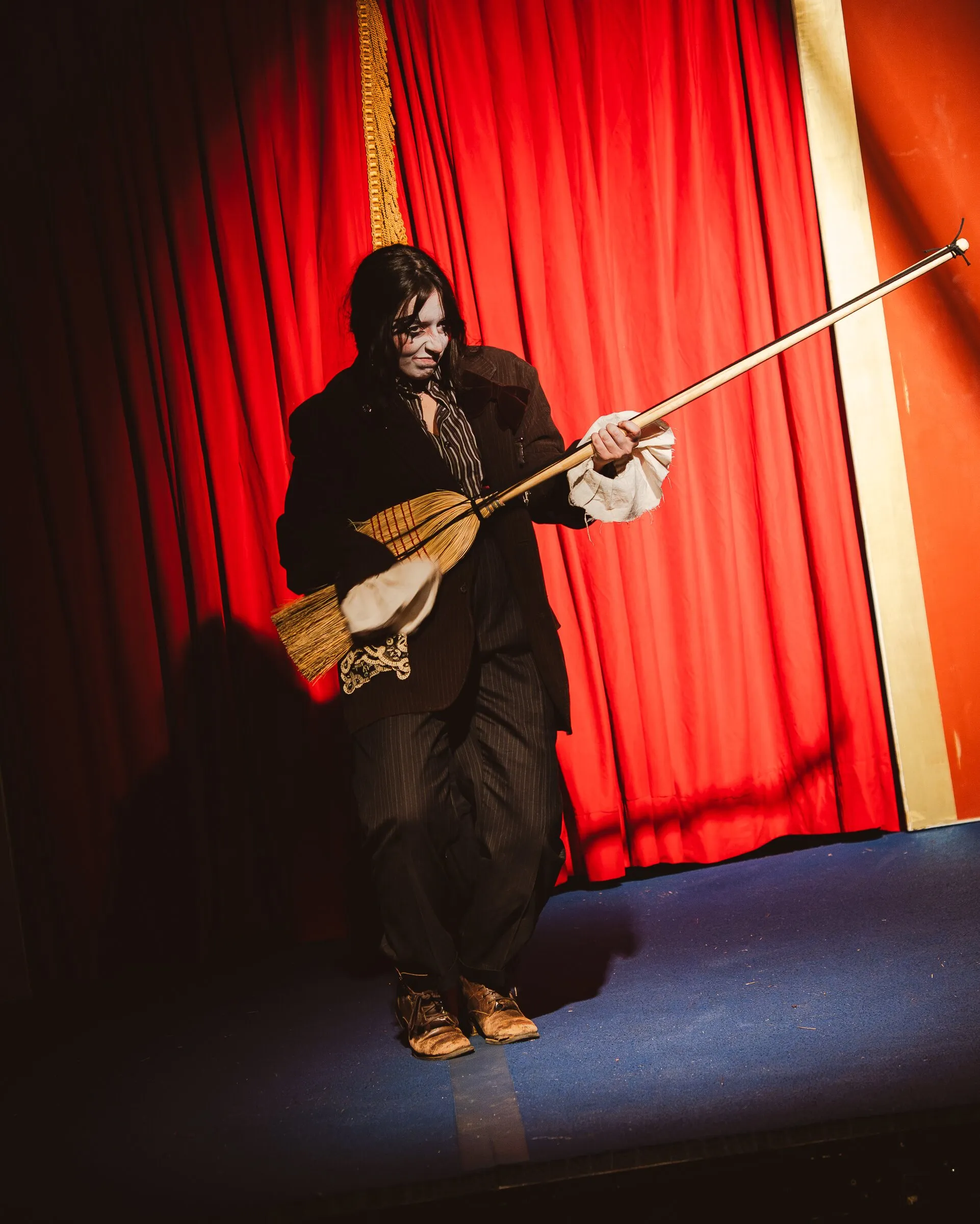A Kuroneko Circus sideshow clown in a shabby suit playing a broom like a fiddle