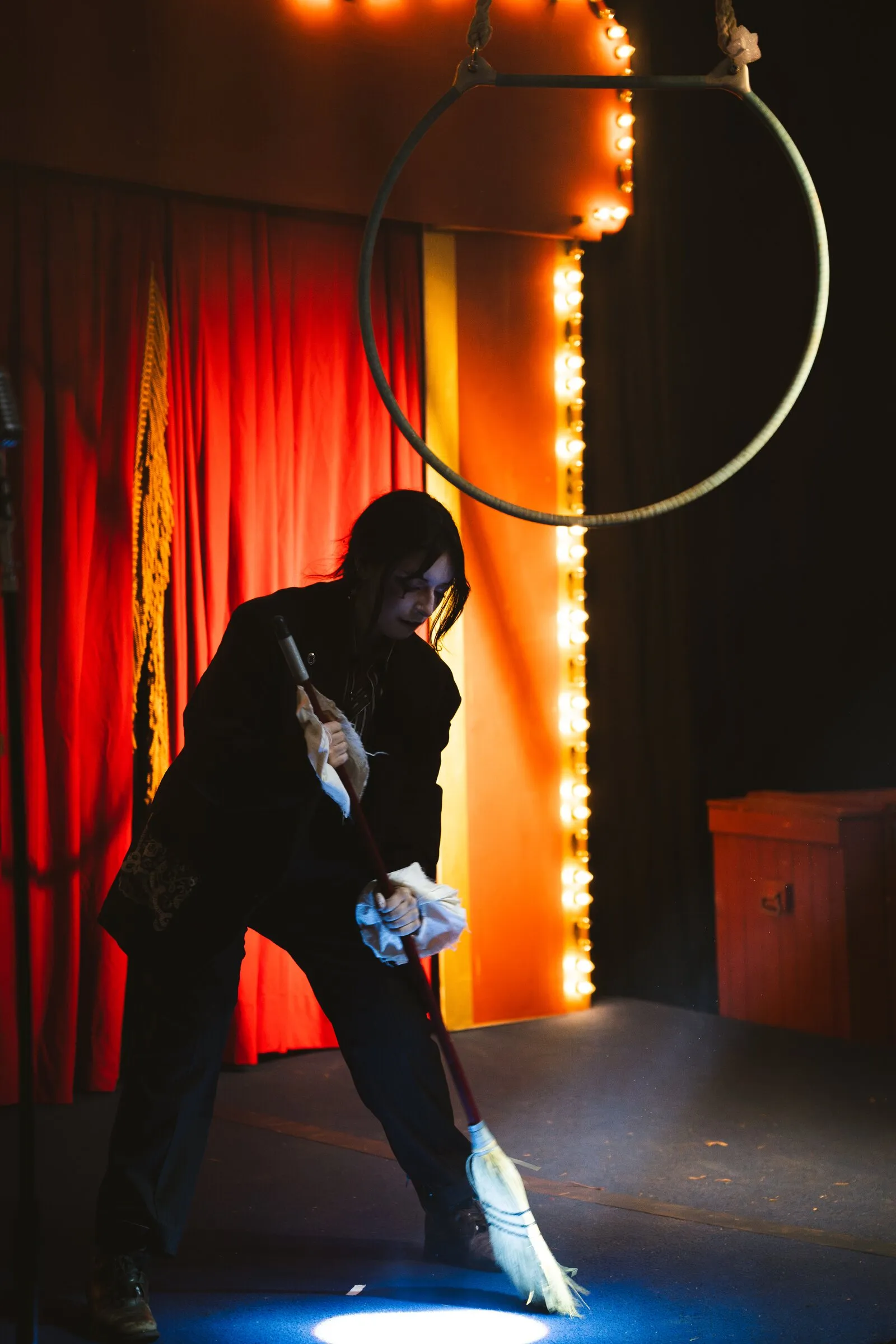 A Kuroneko Circus sideshow clown leaning on a broom in a spotlight beneath an aerial hoop